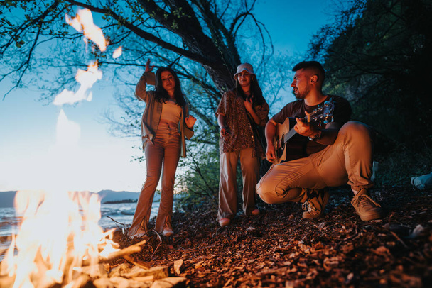 Μια ομάδα φίλων μαζεύονται γύρω από μια ζεστή φωτιά δίπλα στη λίμνη, απολαμβάνοντας τη μουσική που παίζεται σε μια κιθάρα κάτω από τον ουρανό το βράδυ. - Φωτογραφία, εικόνα
