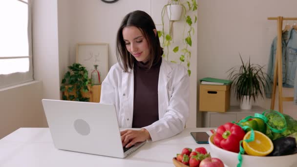 Жіночий дієтолог у білому пальто працює на ноутбуці за офісним столом. Професійна дієтична жінка, яка друкує на клавіатурі, дивлячись на екран комп'ютера на робочому місці. Концепція охорони здоров'я. - Кадри, відео