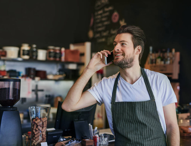 Man, barista and phone call for communication in coffee shop for hospitality, take away and bistro server. Male employee, small business and customer service in cafe for technology and food service. - Photo, Image