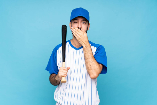 Молодой человек играет в бейсбол на изолированном синем фоне, прикрывая рот руками - Фото, изображение