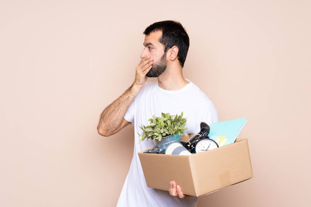 Hombre sosteniendo una caja y moviéndose en un nuevo hogar sobre un fondo aislado cubriendo la boca y mirando hacia un lado - Foto, Imagen