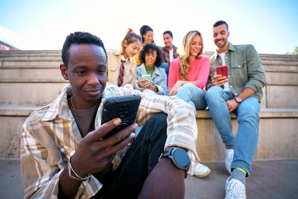 Seriöser Schwarzer mit Smartphone. Afroamerikaner hält Zelle draußen mit Freunden Hintergrund. Unbekümmerte junge Leute in lässiger Kleidung, die gemeinsam Spaß haben. Techniksüchtige Generation - Foto, Bild