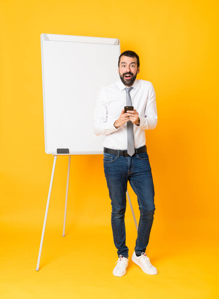 Full-length shot van zakenman geven van een presentatie op wit bord over geïsoleerde gele achtergrond verrast en het verzenden van een bericht - Foto, afbeelding