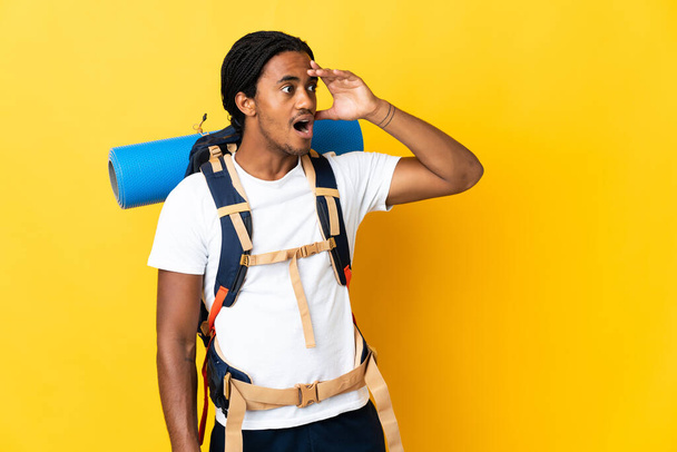 Jonge bergbeklimmer man met vlechten met een grote rugzak geïsoleerd op gele achtergrond doen verrassing gebaar terwijl kijken naar de zijkant - Foto, afbeelding