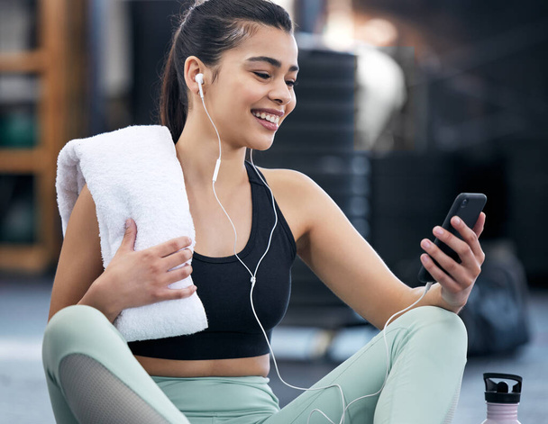 Earphones, gym and woman with phone, watch and video of fitness, exercise and online for wellness. Technology, mobile and social media for happiness, person and athlete on break, relax and towel. - Photo, Image
