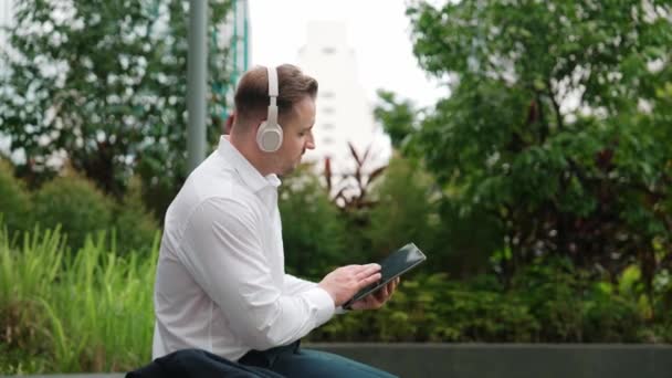 Smart businessman wear headphone to listen relaxed music while take off suit and move to music at green city. Side view of happy project manager with headset dancing movement with lively song. Urbane. - Footage, Video