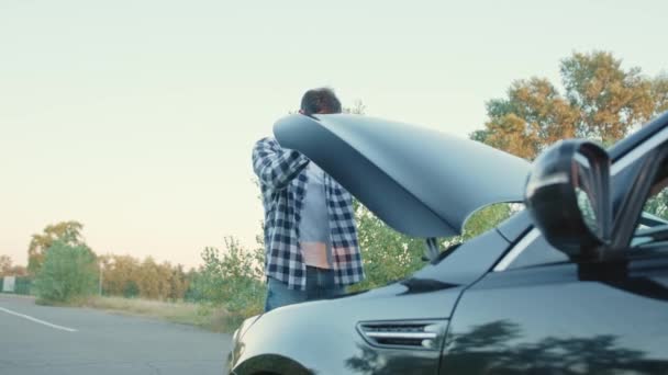 Egy fickó áll egy lerobbant autó mellett, és kinyitja a motorháztetőt a szabadban. Szervizkocsi koncepció. Lassú mozgás. - Felvétel, videó
