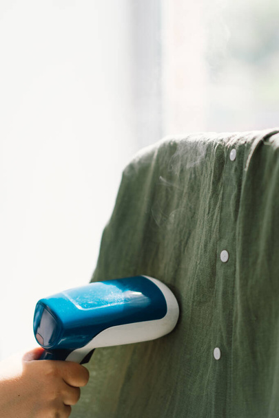 Mujer usando un vapor portátil azul en una prenda verde en casa durante el día. Luz natural que ilumina la textura de las telas y el vapor en acción. - Foto, imagen