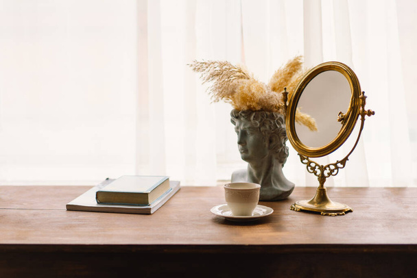 Specchio incorniciato in oro poggia su un tavolo di legno, accanto a un classico vaso busto decorato con pampas erba. Una semplice tazza di caffè bianco completa l'ambiente calmante e sofisticato. - Foto, immagini