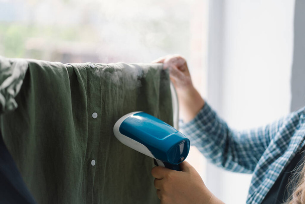 Mulher usando um azul Handheld Steamer em uma roupa verde em casa durante o dia. Luz natural que ilumina a textura dos tecidos e o vapor em ação. - Foto, Imagem