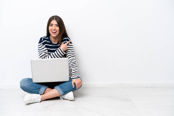 Молода біла жінка з ноутбуком, що сидить на підлозі, ізольована на білому тлі, здивована і спрямована сторона
 - Фото, зображення