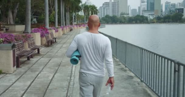 Idősebb férfi sétál a parkban jóga szőnyeg kézzel keresi a tökéletes hely gyakorlás visszapillantás. Man walking city park felfedezése jóga munkamenet reggeli edzés edzés előtt a munka. - Felvétel, videó