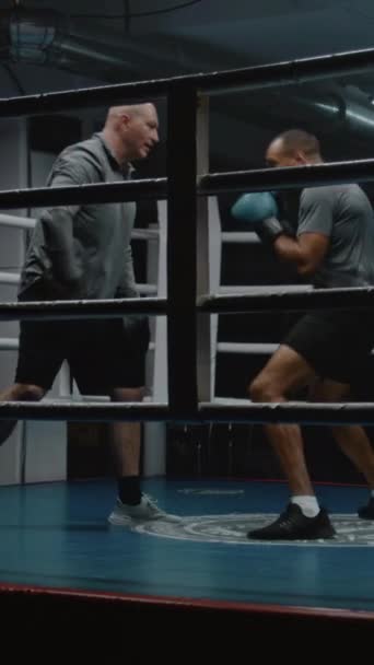 African American man in boxing gloves hits punching mitts and practices in fighting techniques. Athletic boxer prepares for championship with Caucasian coach in dark gym. Vertical shot - Footage, Video
