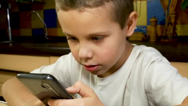 Een schattige jongen van 7-8 jaar zit met een smartphone in zijn handen, communiceert in sociale netwerken. Kind en gadgets. Afstandsonderwijs. Smartphonespelletjes. - Video