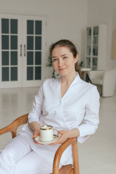Медицина, здравоохранение и люди концепция - Дружелюбная женщина GP врач в белой форме пить кофе чай за завтраком во время работы в офисе клиники. Часть серии. - Фото, изображение
