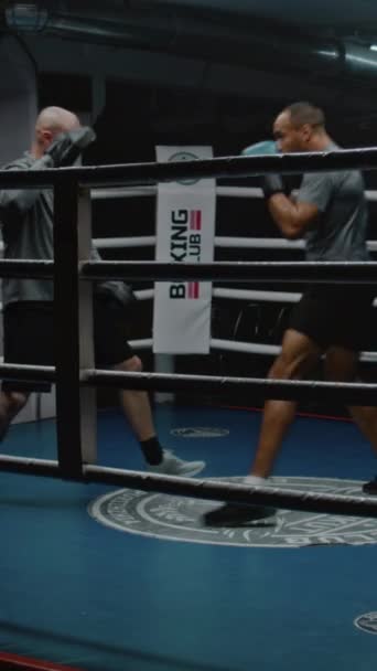 African American fighter exercises and prepares for fighting match with Caucasian trainer on boxing ring. Athletic man in boxing gloves hits punching mitts in dark gym. Vertical shot - Footage, Video