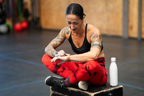 Female athlete sitting on a box using a smart watch after training in a cross training center - Photo, Image
