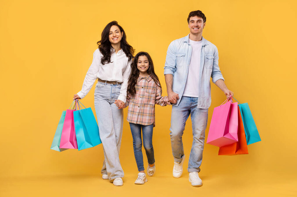 Heureux parents gais et leur fille avec des sacs à provisions lumineux marchant sur fond jaune, profiter des ventes saisonnières et promos - Photo, image