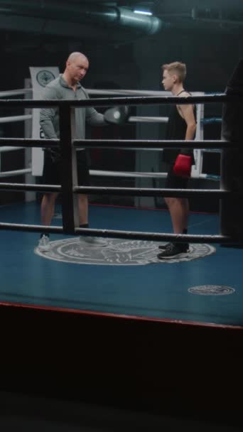 A bokszkesztyűs fiú gyűrűn áll és beszél az edzővel. Felnőtt férfi konzultál fiatal bokszoló és magyarázza harci technikák. A tinédzser harcra készül a sötét edzőteremben. Függőleges lövés - Felvétel, videó