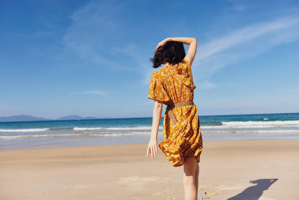 Γυναίκα με κίτρινο φόρεμα στέκεται στην παραλία με την πλάτη στον ωκεανό απολαμβάνοντας ειρηνική στιγμή από την άκρη του νερού - Φωτογραφία, εικόνα