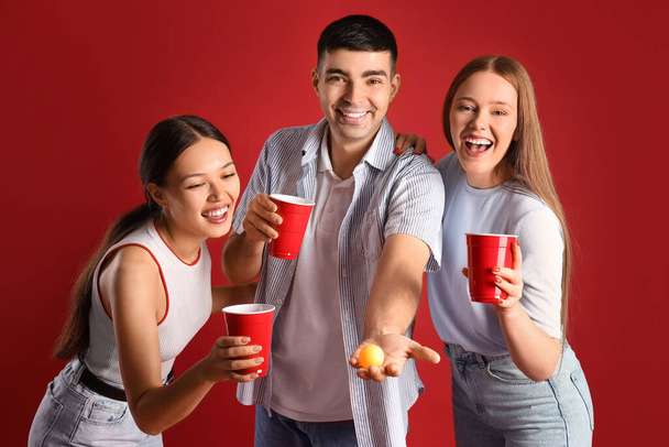 Молодые друзья с чашками и мячом для пиво-понга на красном фоне - Фото, изображение
