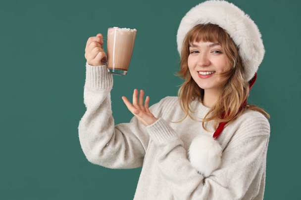 Pretty young woman in Santa hat showing cup of hot chocolate with marshmallows on green background - Photo, Image