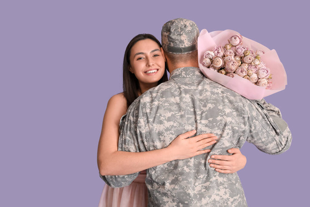Hombre en uniforme militar abrazando a su esposa con flores sobre fondo lila, vista trasera. Celebración de San Valentín - Foto, Imagen