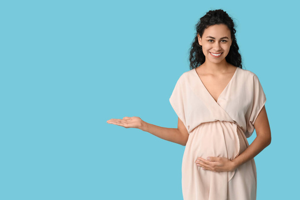 Όμορφη νεαρή έγκυος Αφρο-Αμερικανίδα δείχνει κάτι σε μπλε φόντο - Φωτογραφία, εικόνα