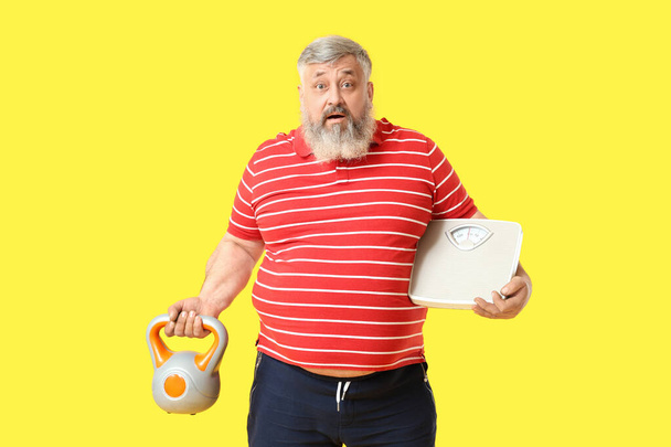 Υπερβολικό βάρος σοκαρισμένος ώριμος άνθρωπος με κλίμακες και kettlebell σε κίτρινο φόντο. Έννοια απώλειας βάρους - Φωτογραφία, εικόνα