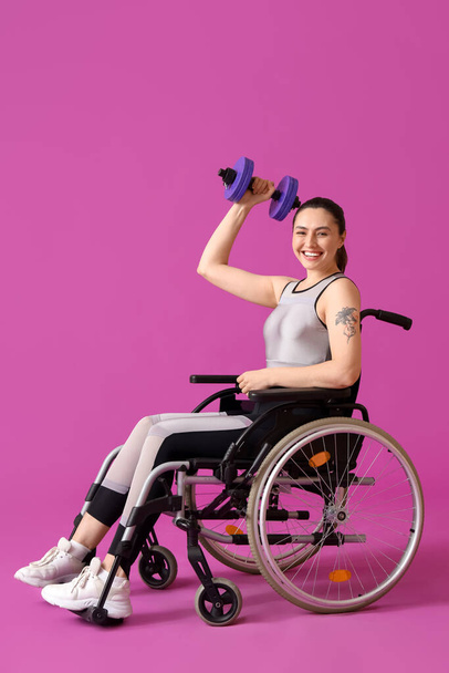 Mujer joven deportiva en silla de ruedas con mancuerna sobre fondo morado - Foto, Imagen