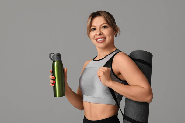 Entrenadora de fitness femenina joven con esterilla de yoga y botella de agua sobre fondo gris - Foto, imagen