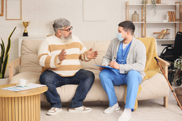 Hombre mayor hablando con el cuidador en la máscara en el sofá en casa - Foto, Imagen