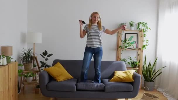 Happy beautiful young woman listening to music with smartphone while dancing on the sofa in the living room at home. Leisure concept. - Footage, Video