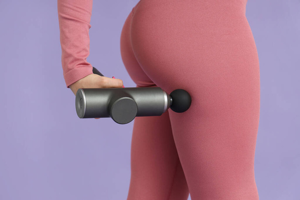 Sporty young woman massaging her thigh with percussive massager on lilac background, closeup - Photo, Image