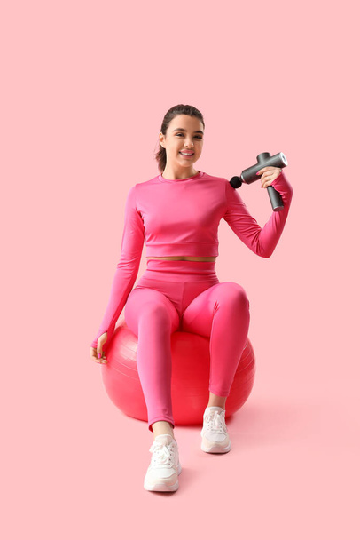 Sporty young woman massaging her shoulder with percussive massager on fitball against pink background - Photo, Image