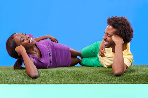 Ευτυχισμένο, μαύρο ζευγάρι και ξαπλωμένος με πράσινο γρασίδι για αγάπη, ρομαντισμό ή υπαίθριο Σαββατοκύριακο με φόντο ένα μπλε στούντιο. Αφρικανός άνδρας και γυναίκα γελάνε χαλαρώνοντας για να διασκεδάσουν οι διακοπές δένονται στο χώρο mockup. - Φωτογραφία, εικόνα