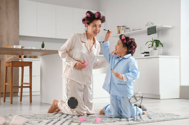 Pequeña chica asiática y su madre con rizadores de pelo aplicando rímel en la cocina - Foto, imagen