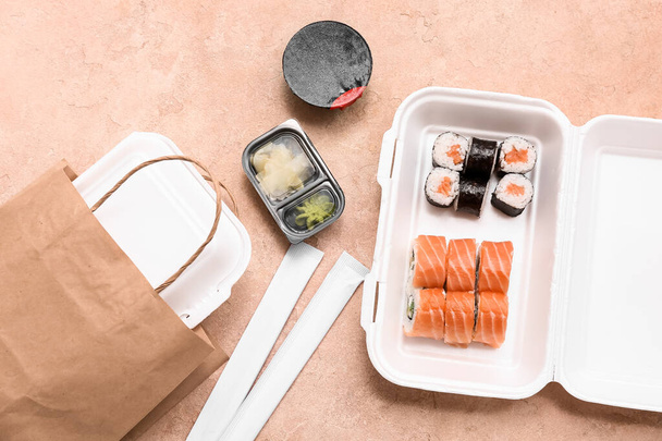 Plastic container with tasty sushi rolls, soy sauce and paper bag on beige grunge background. Delivery concept - Photo, Image