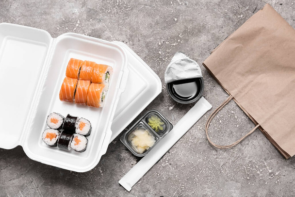 Plastic containers with tasty sushi rolls, chopsticks and paper bag on grey grunge background. Delivery concept - Photo, Image