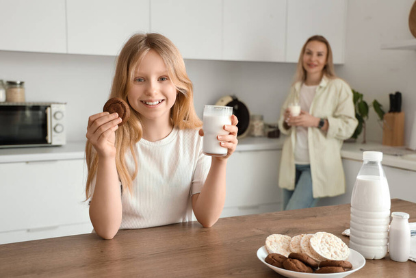 Ευτυχισμένη κόρη και η μητέρα της με κούπες γάλα και νόστιμα μπισκότα στην κουζίνα στο σπίτι - Φωτογραφία, εικόνα