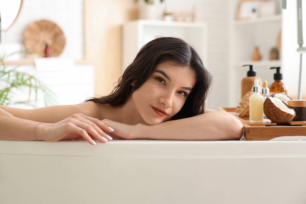 Beautiful young woman with bottles of hair products and coconut in bathroom at home - Photo, Image