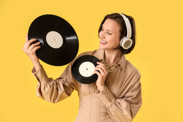 Mooie jonge vrouw in hoofdtelefoon met vinyl schijven op gele achtergrond - Foto, afbeelding