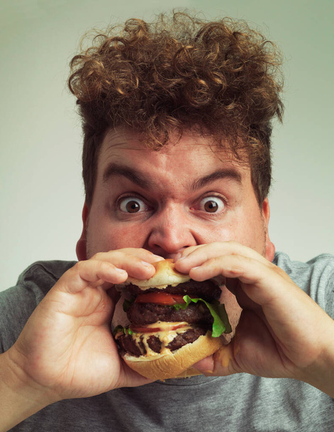 Takeaway, burger and food for man in studio with meal for big eater, plus size or greed. Young person with meat in mouth for hunger, silly and bite indoor for delicious, taste and sauce for portrait. - Photo, Image