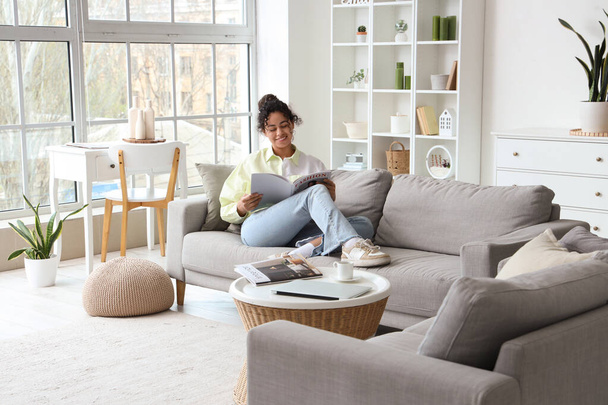 Young African-American woman reading magazine on sofa at home - Photo, Image