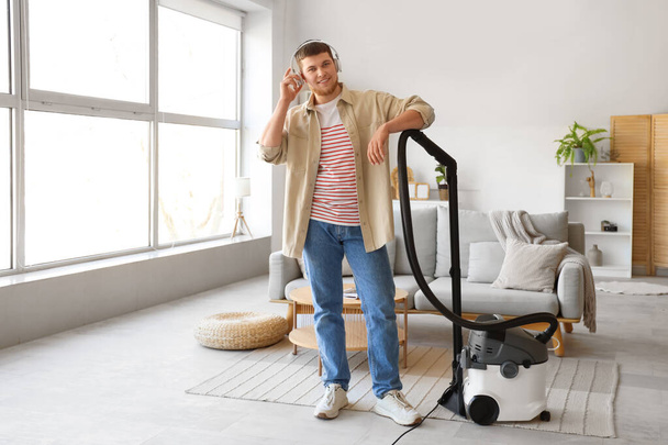 Young bearded man in headphones with vacuum cleaner at home - Foto, imagen