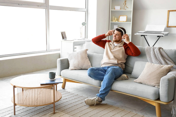 Mladý vousatý muž ve sluchátkách poslouchá hudbu na pohovce doma - Fotografie, Obrázek