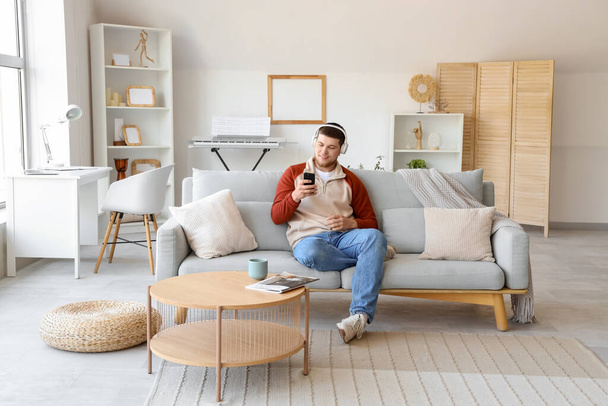 Junger bärtiger Mann mit Kopfhörer und Handy hört zu Hause auf dem Sofa Musik - Foto, Bild