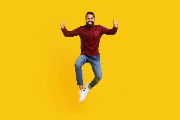Indiase man is gevangen midden in de lucht, springen energiek met zijn handen opgeheven in duim ups gebaar, gele studio achtergrond - Foto, afbeelding