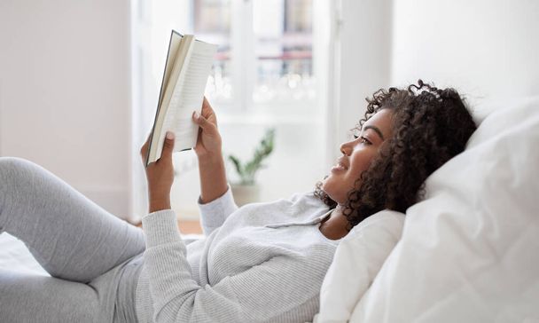 A Hispanic woman is lying down in bed with a book in hand, engrossed in reading. The room is dimly lit, and she appears relaxed and focused on the pages of the book, side view - Photo, Image