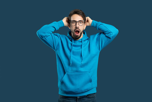 Egy fiatal férfi kék kapucnis pulóvert és szemüveget visel, nyitott szájjal áll, kezeit a fejére helyezi a tiszta kék háttér előtt, meglepetését vagy hitetlenségét fejezve ki váratlan helyzetekben.. - Fotó, kép